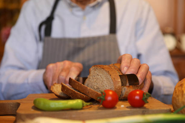 An older man is cooking lunch and preparing bread in the kitchen. The scene captures him in a close-up as he focuses on his tasks. The kitchen background features various utensils, ingredients, and appliances, creating a busy and productive atmosphere.\