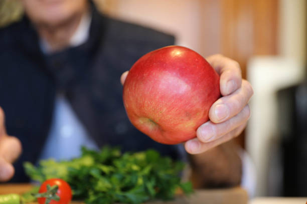 An elderly man is holding an apple in his hand, with a close-up focus. The image captures the texture and details of the apple and the man’s hand, emphasizing the fruit's freshness and the man's gentle grip. The background is softly blurred, highlighting the apple and the man's hand