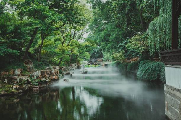 A tranquil Chinese garden with mist rising from a still pond, surrounded by lush greenery and a stone bridge.