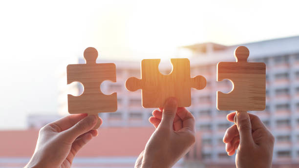 istock Teamwork and partnership concept. Hands are raised against a bright, white sky, each holding a piece of a wooden jigsaw puzzle. teamwork, problem-solving, Charity, volunteer. Unity, team business. 2164750704