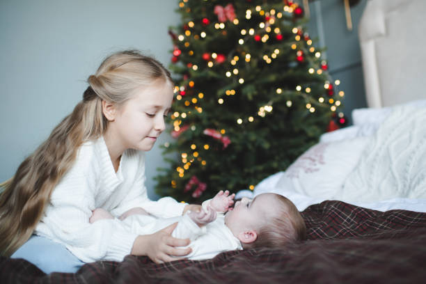Happy child girls playing in bed together over Christmas glowing tree at home in room close up. Winter holiday season. Childhood. Celebration.