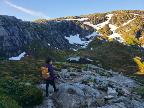 A lone hiker traverses rocky terrain, surrounded by towering mountains and patches of snow, basking in the sunlight. Lysefjord Kjeragbolten Kjerag Norway