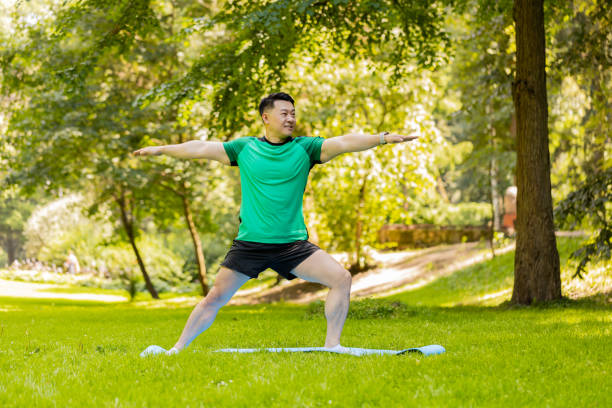 istock Asian man performs gymnastics stretching practicing yoga warrior pose on mat in urban city park 2164801727