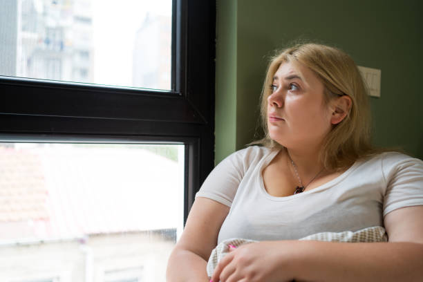 istock Plus size woman sitting alone by window, looking outside with thoughtful expression. Depression, mental health issues, heartbreak, or financial difficulties 2164817915