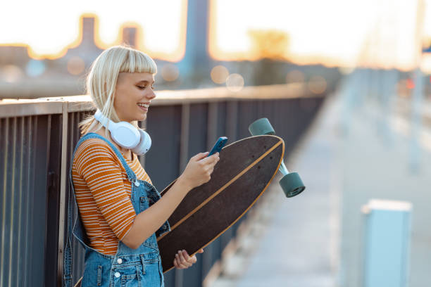 Young urban woman holding longboard and using smart phone