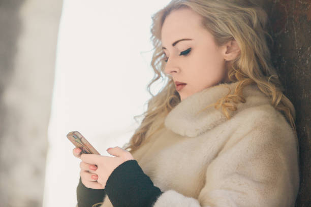 Young woman with blonde hair and a 1950s vintage style, wearing a white fur coat, is using a smartphone in a european city