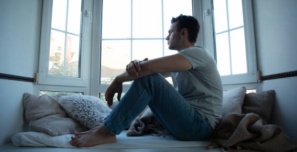 istock Depressed, sad, frustrated, crying, scared, instability mental and bipolar young man sitting on the windowsill at home. Headache because overworked and stress 2164855803