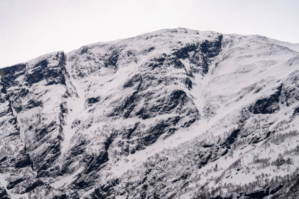 A breathtaking view of a snow-capped mountain cliff rising steeply against the clear sky. The rugged rock formations and patches of snow create a stunning contrast, showcasing the raw beauty and grandeur of the mountainous landscape