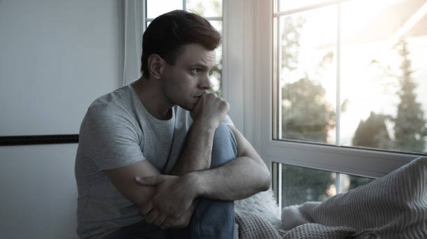 istock Depressed, sad, frustrated, crying, scared, instability mental and bipolar young man sitting on the windowsill at home. Headache because overworked and stress 2164856080