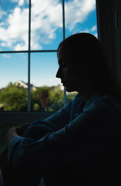 istock Vertical image of shadowed silhouette depressed woman while she sitting on windowsill with wide window on background. Sad, crisis, bad emotions, mental health, family violence. 2164856104