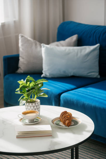 Side view of coffee cup with espresso close to croissant and books on a white table in front of a blue sofa. Weekend in hotel, comfortable living room concepts