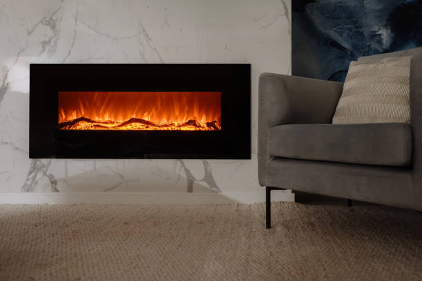 Living room with electric fireplace and chair against marble wall Wide shot of modern electric fireplace with realistic flames mounted on white marble wall. Cozy gray chair sits near fireplace in living room. Quiet evening in hotel or weekend at home modern cottage interior design stock pictures, royalty-free photos & images