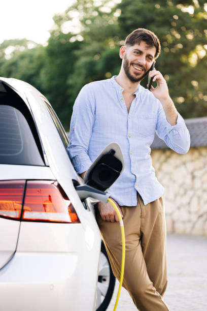 istock Portrait of handsome joyful 30-aged man with beard which has mobile conversation while luxurious car charging battery on specially equipped charging station 2164903963