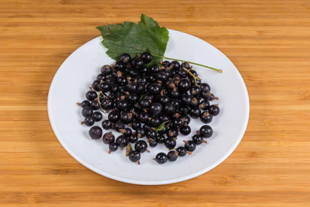 Ripe freshly harvested blackcurrant with green leaf on the white dish on a wooden surface