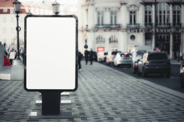Blank advertising billboard on a busy city street. The billboard stands on a tiled pavement with cars and historic buildings in the background, ready for customized promotional content
