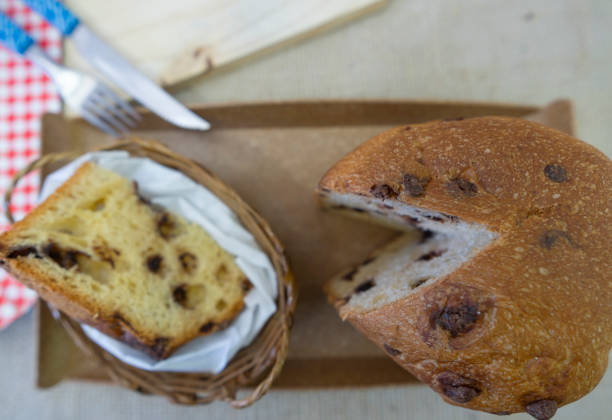 Stock photo showing close-up view of a traditional, dome-shaped panettone, an Italian sweet bread, decorated with a pinecone being dusted with sifted icing sugar, displayed on a china cake stand with the base surrounded by variegated holly leaves and red berries. The fruit cake is being dusted by sifted icing sugar.