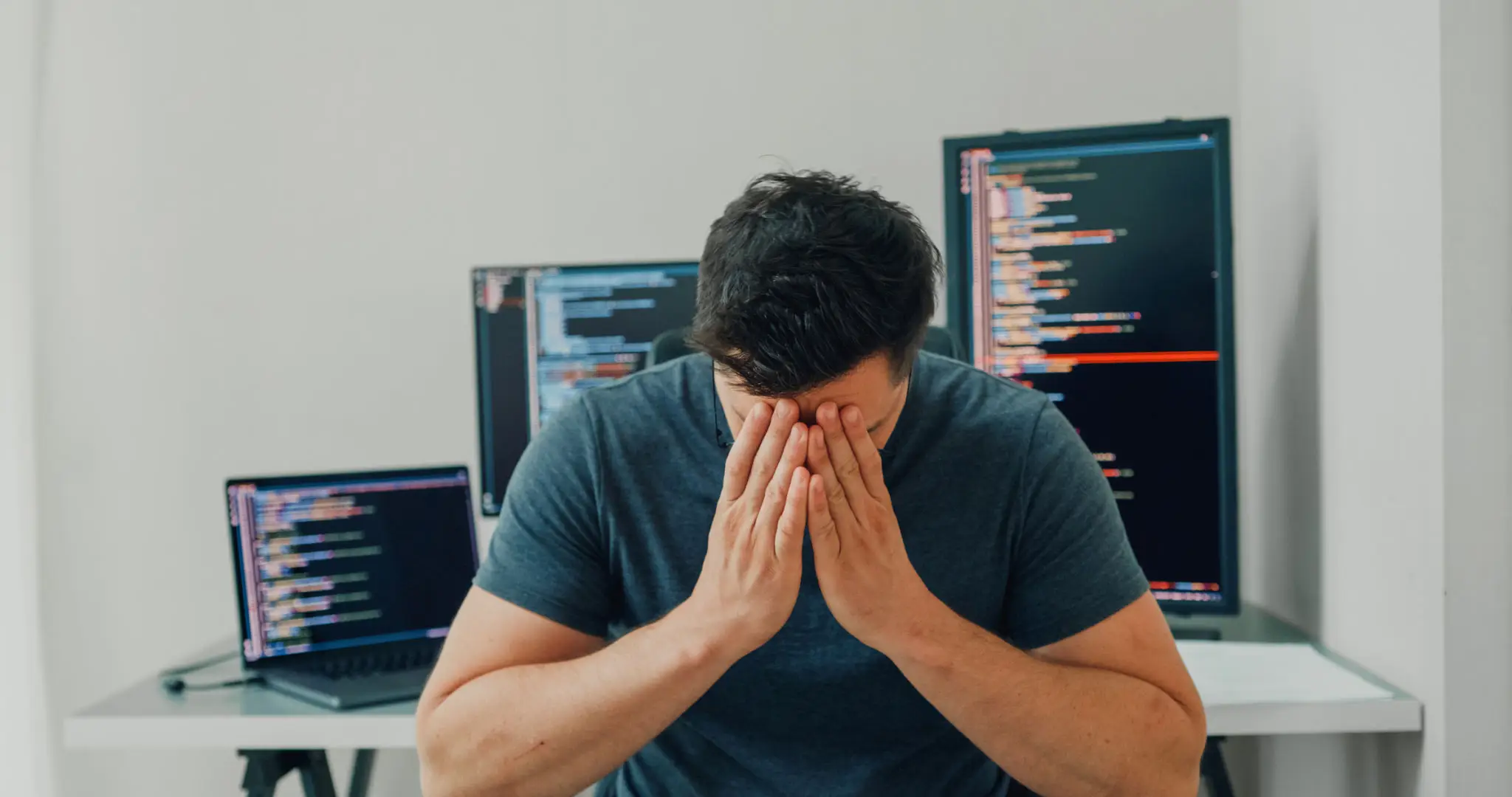 Tired depressed bored man software developers for AI technology looking at camera sitting at desk with multiple screens work at home office. Programmer development.