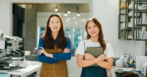 Two happy female entrepreneurs, a mentor and her trainee, pose with confidence at their successful and sustainable coffee shop. Small business owner.