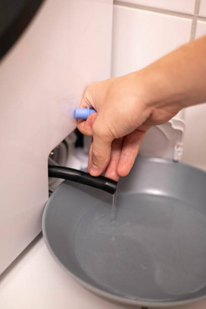 istock A hand drains water from a washing machine using a small hose. The water flows into a bowl, showing a maintenance task. This step helps ensure the appliance's efficiency and longevity. 2164993180