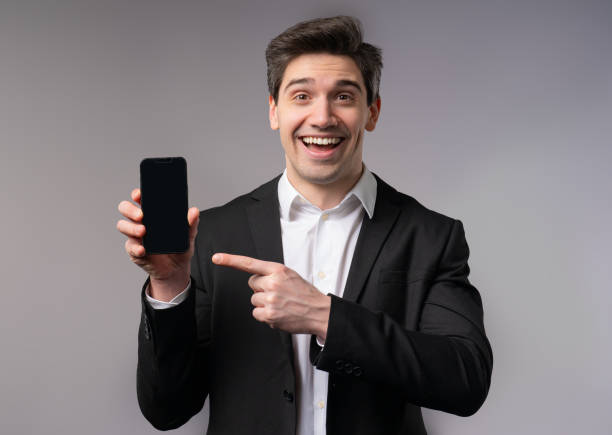 istock Handsome businessman pointing with finger on mobile phone screen on grey studio wall. Modern technology, copy space. 2164993249