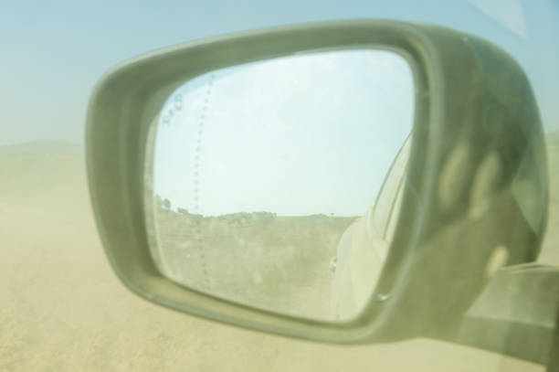 A reflective rearview mirror captures a long winding road disappearing into the horizon with the silhouette of distant mountains, hinting at a journey ahead on a sunny day.