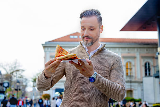 istock Young man eating pizza while walking through a city 2165015679