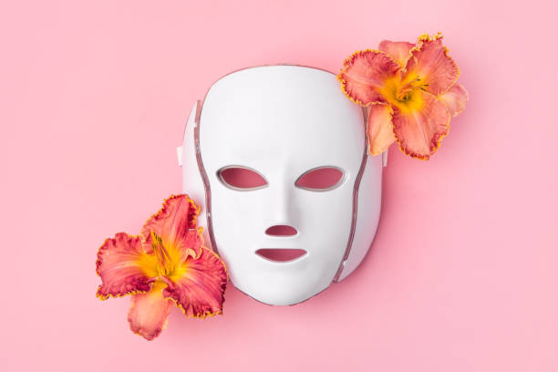 LED facial mask Cosmetic LED facial mask regenerative treatment on pink background with flowers lilies. Health and beauty of the face. mask stock pictures, royalty-free photos & images