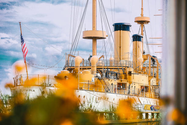Philadelphia, Pennsylvania, July 19, 2024. A historic ship with the name Olympia is docked, adorned with towering funnels and an American flag on its stern. Foreground elements of blurry flowers add a contrast to the towering structure of the vessel.