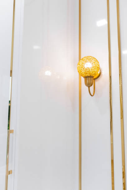 classic-style wall sconce that exudes luxury and elegance. The sconce is crafted from shiny gold material, creating a sophisticated and upscale aesthetic. It is mounted on a clean white wall, which enhances its prominence and visual appeal.
