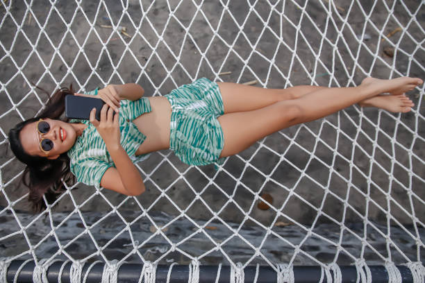 Directly above shot of Indonesian woman using phone on a hanging suspended net hammock at a tourist resort by the beach.concept: travel influencer making content ,relaxing on hammock, selfie on hammock.checking social media, telecommuting on the beach
