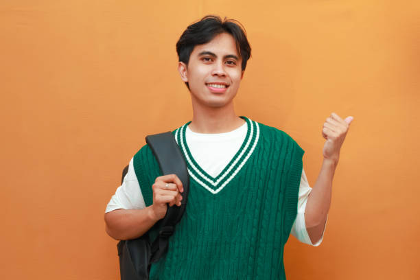 istock Happy Asian student wearing green sweater posing while carrying backpack and pointing to the side with thumbs up while smiling friendly isolated on orange background 2165045597