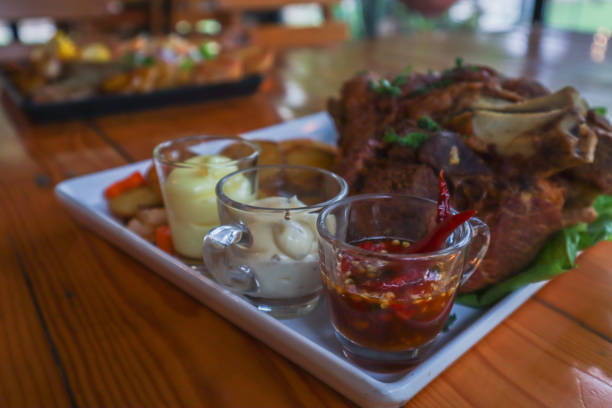 German pork knuckle is served with baked potatoes and sauce inside a white ceramic plate placed on the dining room table to make the pork knuckle ready for a dinner feast.