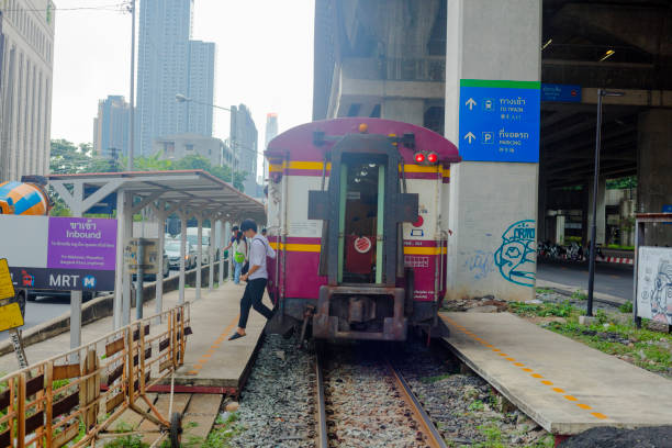 Back of old thai train at Makkasan station in Bangkok. Incoming old train has just stopped at Makkasan railway station below modern airport railway link station at daytime with overcast. Train is old and last wagon is purple colored. People are exiting train to platform at Kampheng Phet Road. A young man is stepping out of train and last wagon.  In background is skyline of Bangkok Pathum Wan downtown area.
