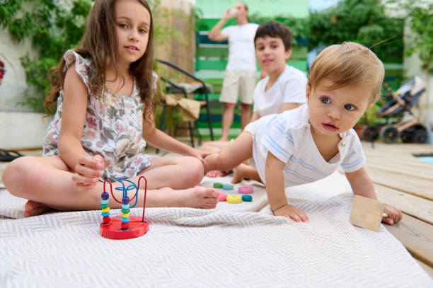 Children playing with toys outside on a sunny day while their father watches in the backyard Children enjoying playtime with toys outdoors on a sunny day in their backyard with their father observing in the background. 6-11-months stock pictures, royalty-free photos & images
