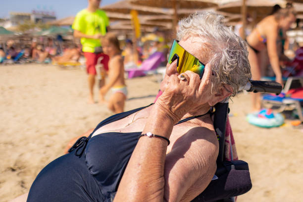 An older woman, covered with a beach towel, sits in her wheelchair gazing at the calm sea.