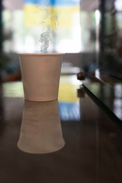 A paper disposable cup with a hot drink on a glass bar counter. Steam is coming out of the cup. Close-up. Vertical photo. In the background is a part of the Ukrainian flag. High quality photo