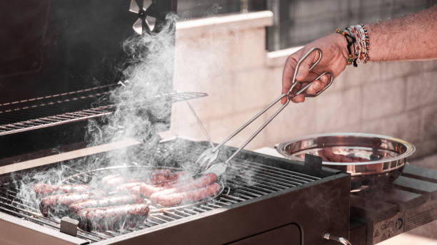Man's hands grabbing with long tongs the meats he is cooking on the BBQ. Panoramic format and dark edition.