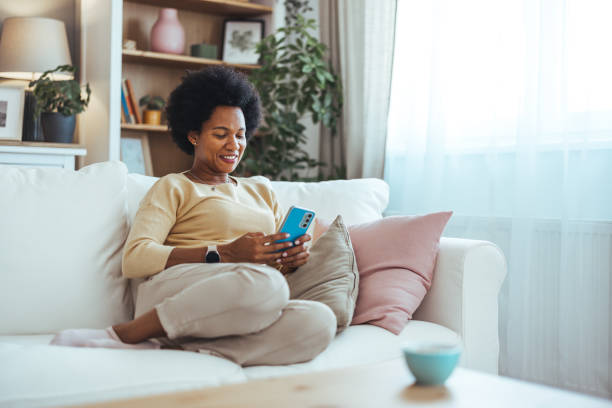 istock A smiling elegant African-American female using her smartphone while sitting on the cozy sofa in the living room. Beautiful mixed race woman browsing internet on cellphone in home living room. 2165152600