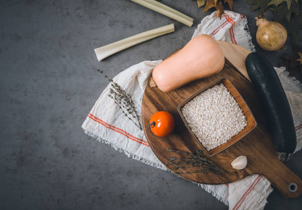 A set of autumn vegetables and rice for making risotto lie on a wooden board with a kitchen napkin on the right against a dark gray background with space for text on the left, top view close-up.