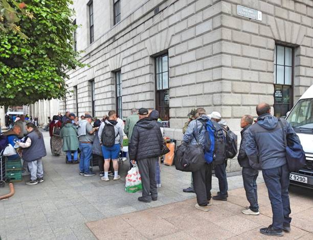 People with backpacks and bags standing in line outside a stone building, likely a food bank or shelter.