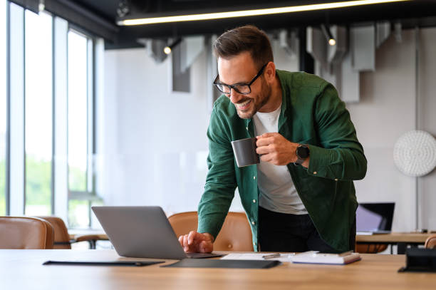 istock Smiling male freelancer drinking coffee and checking e-mails over wireless computer on desk in office 2165225079