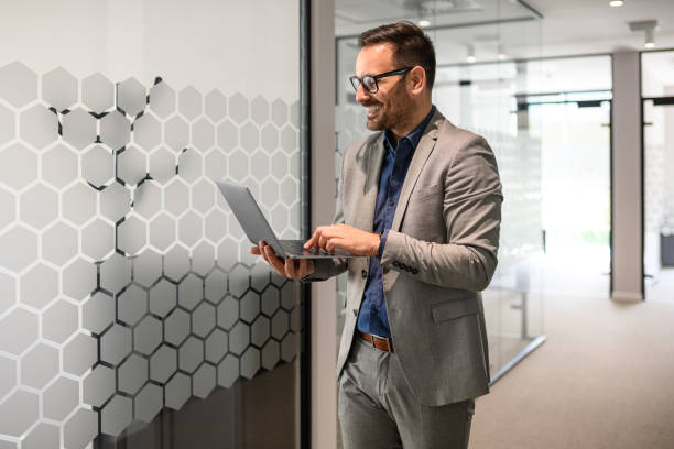 istock Male manager in glasses smiling and working on project over laptop while standing in office corridor 2165225172