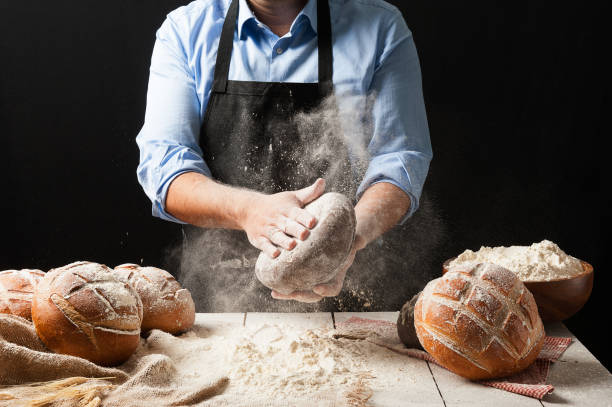 istock Male baker wearing black apron holding a freshly baked rustic bread splashing flour in front of a white wooden table filled with different kinds of loaves and jutte fabric 2165226064