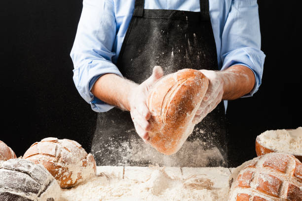 istock Male baker wearing black apron holding a freshly baked rustic bread splashing flour in front of a white wooden table filled with different kinds of loaves and jutte fabric 2165226100