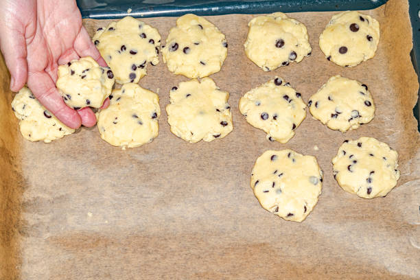 White dough and chocolate chips on a baking tray covered with baking paper. Overhead personal point of view.
