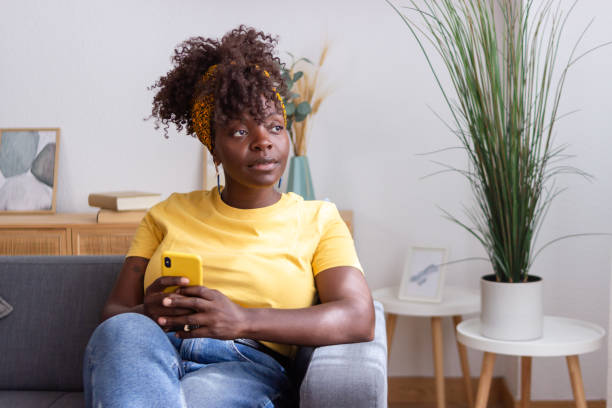istock Young woman holding smartphone is thinking while sitting on sofa at home 2165237726