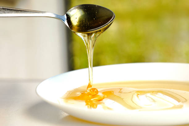Thick, fresh honey is poured in a thin stream from a teaspoon into a white plate. Golden, aromatic honey spreads throughout the plate.