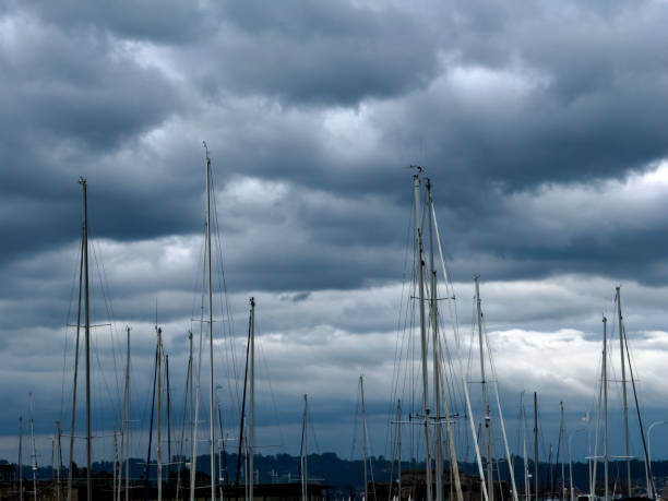 The tall masts of sailboats stand prominently against a dramatic, cloudy sky in a marina. The overcast weather and clustered masts create a striking contrast, capturing the essence of a calm day by the harbor. The scene evokes a sense of tranquility and anticipation as the boats await their next journey.