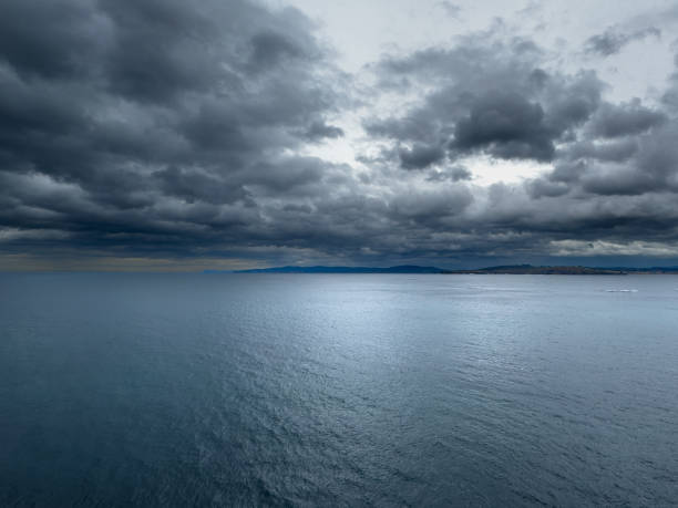 A calm ocean is captured under an overcast sky filled with dark, heavy clouds. The horizon meets the distant mountains, blending seamlessly with the moody sky. This tranquil seascape evokes a sense of calm and quietness, highlighting the beauty of nature before a potential storm.