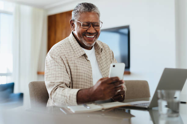 Happy mature male using phone for connecting and staying in touch Happy mature male using his phone while sitting at a desk, connecting and staying in touch. The setting appears comfortable and modern. black man laptop stock pictures, royalty-free photos & images
